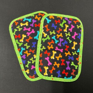 May include: Two rectangular dog bone-themed pot holders. The pot holders have a black background with colorful bones in red, orange, yellow, green, blue, and purple. They are outlined with a bright green trim.