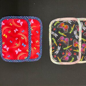 May include: Two square fabric pot holders. One is red with butterflies and curved lines, trimmed with blue and white rope-like trim. The other is black with butterflies, dragonflies, and grasshoppers, trimmed with a patterned border.