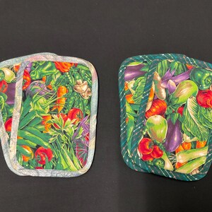 May include: Two square pot holders with a colorful vegetable print. The design features tomatoes, carrots, eggplants, and green peppers. The edges are trimmed with a patterned fabric. The background is black.