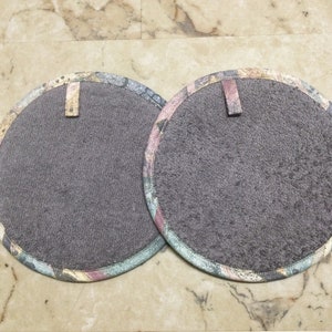 May include: Two round, gray fabric pot holders with a decorative patterned border in shades of pink, blue, and gold. Each pot holder has a small fabric loop for hanging. The pot holders are approximately 8 inches in diameter.