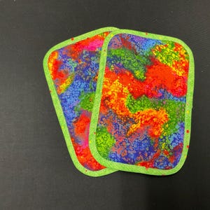 May include: Two rectangular fabric coasters with rounded corners. The coasters feature a vibrant, colorful pattern of red, orange, yellow, green, blue, and purple. Each coaster is outlined with a bright green border.