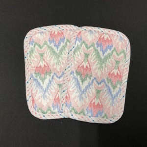 May include: Two square pot holders with a colorful zigzag pattern in pink, green, and blue on a white background. The edges are trimmed with a dotted border. The pot holders are designed for kitchen use.