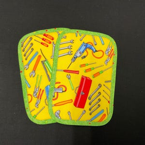 May include: Two yellow potholders with a green trim. The potholders are decorated with a pattern of various tools, including wrenches, screwdrivers, and a drill. A red toolbox is also featured in the design.