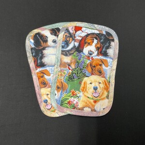 May include: Two matching pot holders with a colorful dog-themed print. The design features various dog breeds, including golden retrievers, dachshunds, and beagles, set against a floral and garden backdrop. The pot holders have a quilted appearance.
