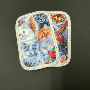 May include: Two square pot holders with a colorful cat print. The fabric features various cat breeds in shades of orange, gray, white, and black, surrounded by flowers. The edges are trimmed with a white and red dotted pattern.
