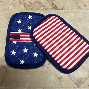 May include: Two blue and white pot holders with an American flag design. One pot holder has a red, white, and blue striped design, and the other has a blue background with white stars and a red, white, and blue stripe.