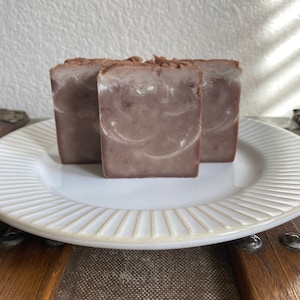 May include: Three bars of handmade soap with a brown and white swirl pattern. The soap is on a white plate with a decorative rim.
