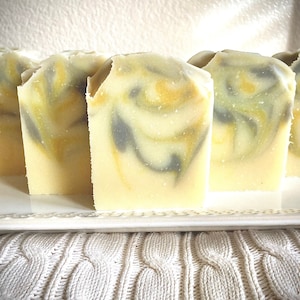May include: Four bars of handmade soap with a swirl pattern of yellow and grey. The soap bars are arranged in a row on a white plate.