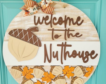 May include: A round wooden door hanger with a white and brown background. The words "Welcome to the Nuthouse" are written in brown letters. The door hanger is decorated with brown acorns and orange leaves.