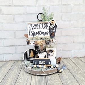 May include: A two-tiered wooden tray with a black and white farmhouse Christmas theme. The top tier features a sign that reads "Farmhouse Christmas" and a small wooden house with a wreath. The bottom tier has a black and white patterned circle, a small wooden tree, and a wooden sign with a bow. The tray is decorated with a black and white gingham ribbon and a beaded garland.