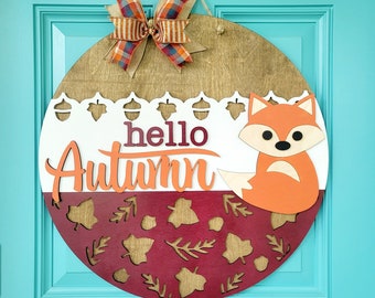 May include: A round wooden door hanger with a white and burgundy background. The top half of the hanger features a white background with a lace trim, the text "hello autumn" and a cutout of an orange fox. The bottom half of the hanger features a burgundy background with a cutout pattern of leaves and acorns. A plaid bow is attached to the top of the hanger.