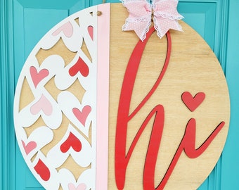 May include: A round wooden door hanger with a pink and white heart pattern on one side and the word "hi" in red on the other side. A pink bow with white polka dots is attached to the top.