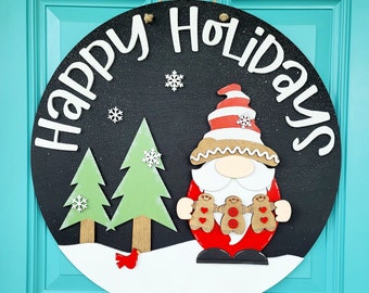 May include: A round wooden door hanger with a black background and white lettering that says "Happy Holidays". A white and red gnome with a white beard and a red hat is holding three gingerbread men. Two green Christmas trees are on either side of the gnome.