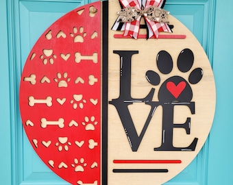 May include: A round wooden door hanger with a red and white paw print pattern on one side and the word "LOVE" with a black paw print on the other side. A red heart is inside the paw print. A red and white gingham bow is at the top.