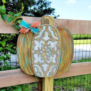 May include: A wooden pumpkin with a carved design painted in white, teal, and orange. The pumpkin is hanging on a wooden fence with a green vine growing on it.