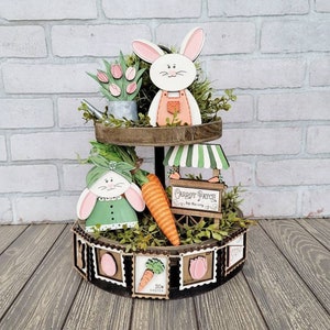 Peut inclure: Un plateau à étages en bois à deux niveaux décoré sur le thème du potager de carottes. Le niveau supérieur présente un lapin blanc aux oreilles roses et une salopette, un arrosoir vert et des tulipes roses. Le niveau inférieur présente un lapin vert avec un nœud rose, une grosse carotte orange et une pancarte qui dit "Carrot Patch Are the Way". Le plateau est décoré d'une guirlande de timbres en bois avec des images de carottes, de tulipes et d'œufs de Pâques.