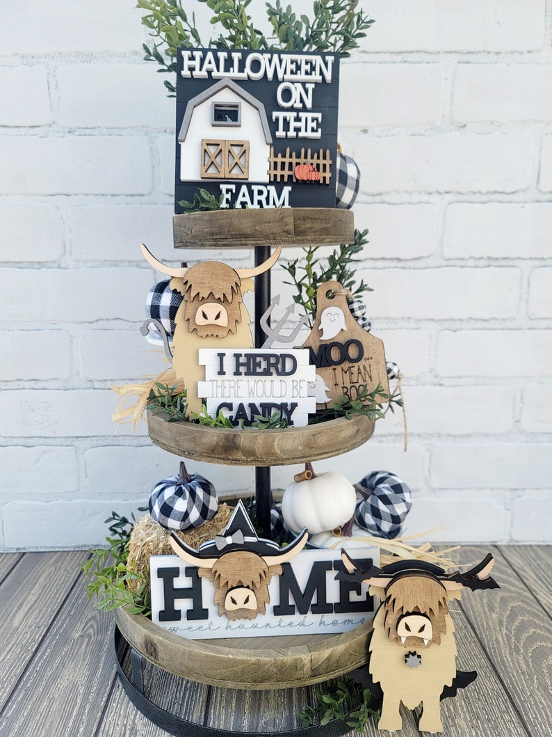 May include: A three-tiered wooden tray decorated for Halloween with a black and white farmhouse theme. The top tier features a sign that says "Halloween on the Farm" with a small barn and pumpkins. The middle tier has a sign that says "I herd there would be candy" and a sign that says "Moo I mean Boo". The bottom tier has a sign that says "Home sweet haunted home" with two Highland cow figurines.