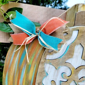 May include: A wooden pumpkin door hanger with a teal, orange, and brown bow. The pumpkin is painted with a multi-colored, abstract design.