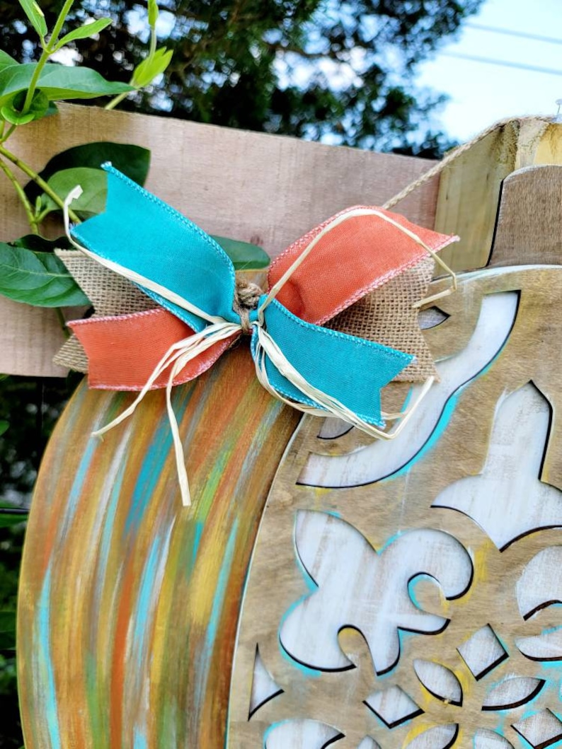 May include: A wooden pumpkin door hanger with a teal, orange, and brown bow. The pumpkin is painted with a multi-colored, abstract design.