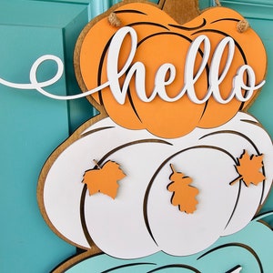 Op de afbeelding: Een houten deurhanger met een gelaagd ontwerp met drie pompoenen. De bovenste pompoen is oranje met het woord "hello" in witte letters. De middelste pompoen is wit met oranje bladeren. De onderste pompoen is teal met het woord "fall" in witte letters.