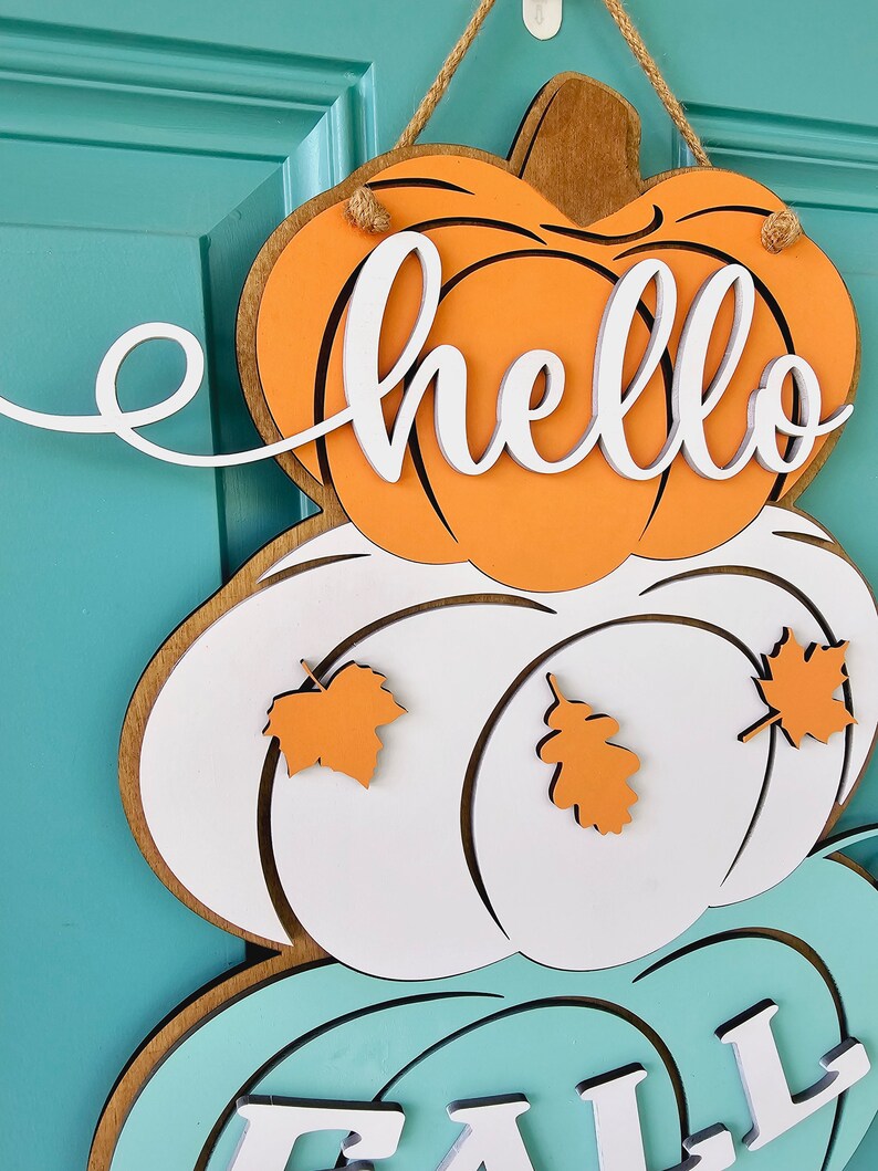 Op de afbeelding: Een houten deurhanger met een gelaagd ontwerp met drie pompoenen. De bovenste pompoen is oranje met het woord "hello" in witte letters. De middelste pompoen is wit met oranje bladeren. De onderste pompoen is teal met het woord "fall" in witte letters.