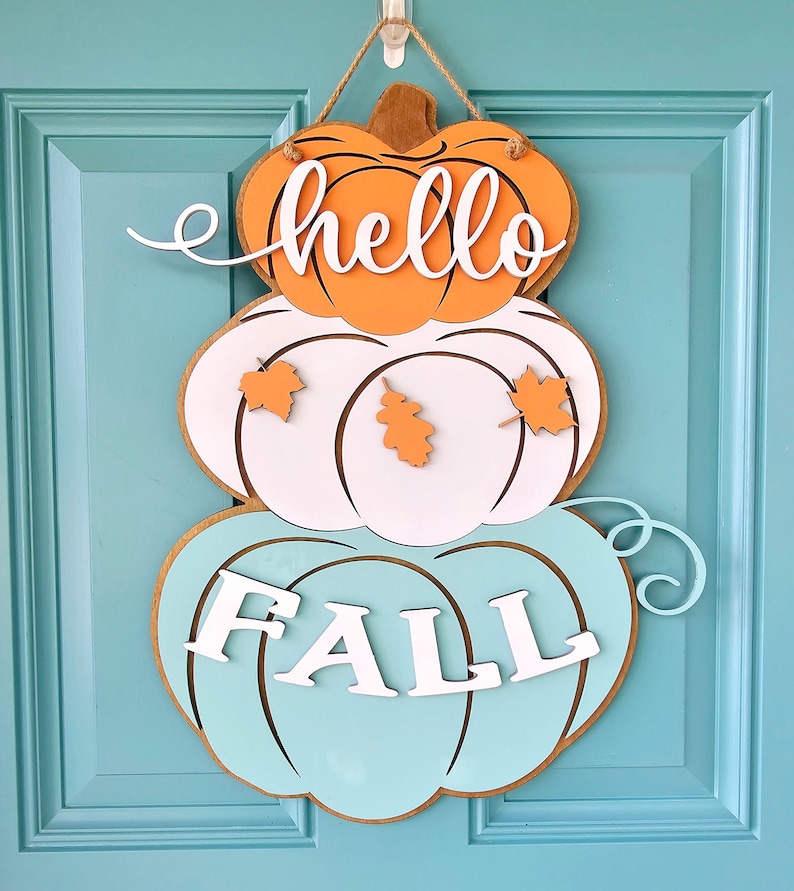 May include: A wooden pumpkin door hanger with three stacked pumpkins. The top pumpkin is orange with the word 'hello' in white letters. The middle pumpkin is white with three orange leaves. The bottom pumpkin is light blue with the word 'FALL' in white letters.