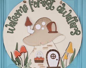 May include: A round wooden sign with the text "Welcome Forest Dwellers" in green lettering. The sign features a brown mushroom with a white roof and a small door. There are also small red and yellow mushrooms on the sign.