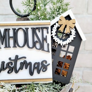 May include: Black and white farmhouse Christmas sign with the words "Farmhouse Christmas" and a small black and white house with a brown bow and a white wreath.