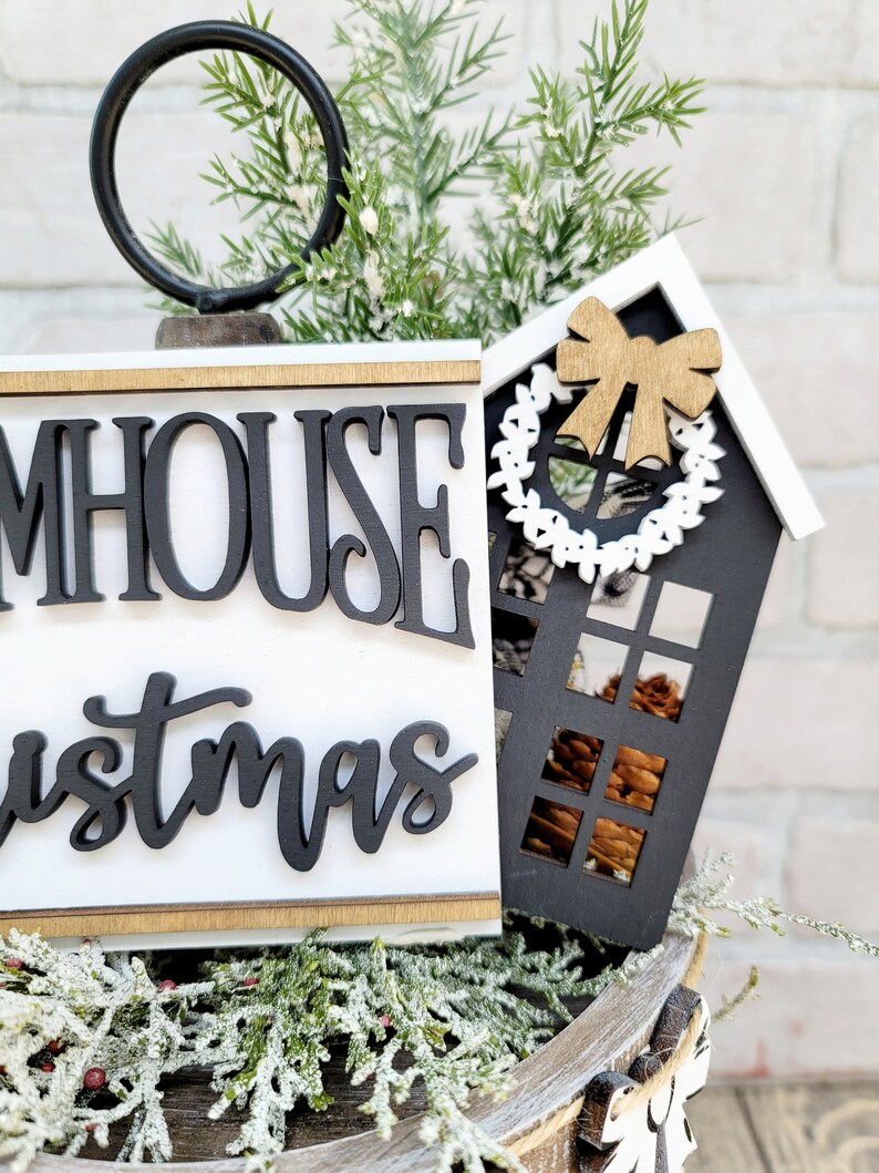 May include: Black and white farmhouse Christmas sign with the words "Farmhouse Christmas" and a small black and white house with a brown bow and a white wreath.