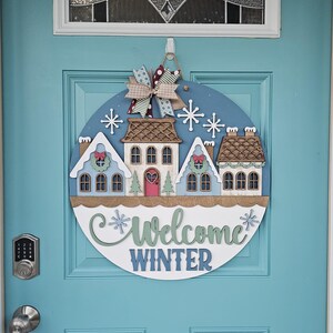 May include: A round wooden door sign with a winter theme. It features a blue background with white snowflakes and a row of houses. The sign says "Welcome Winter" in green and blue. A decorative bow hangs at the top.