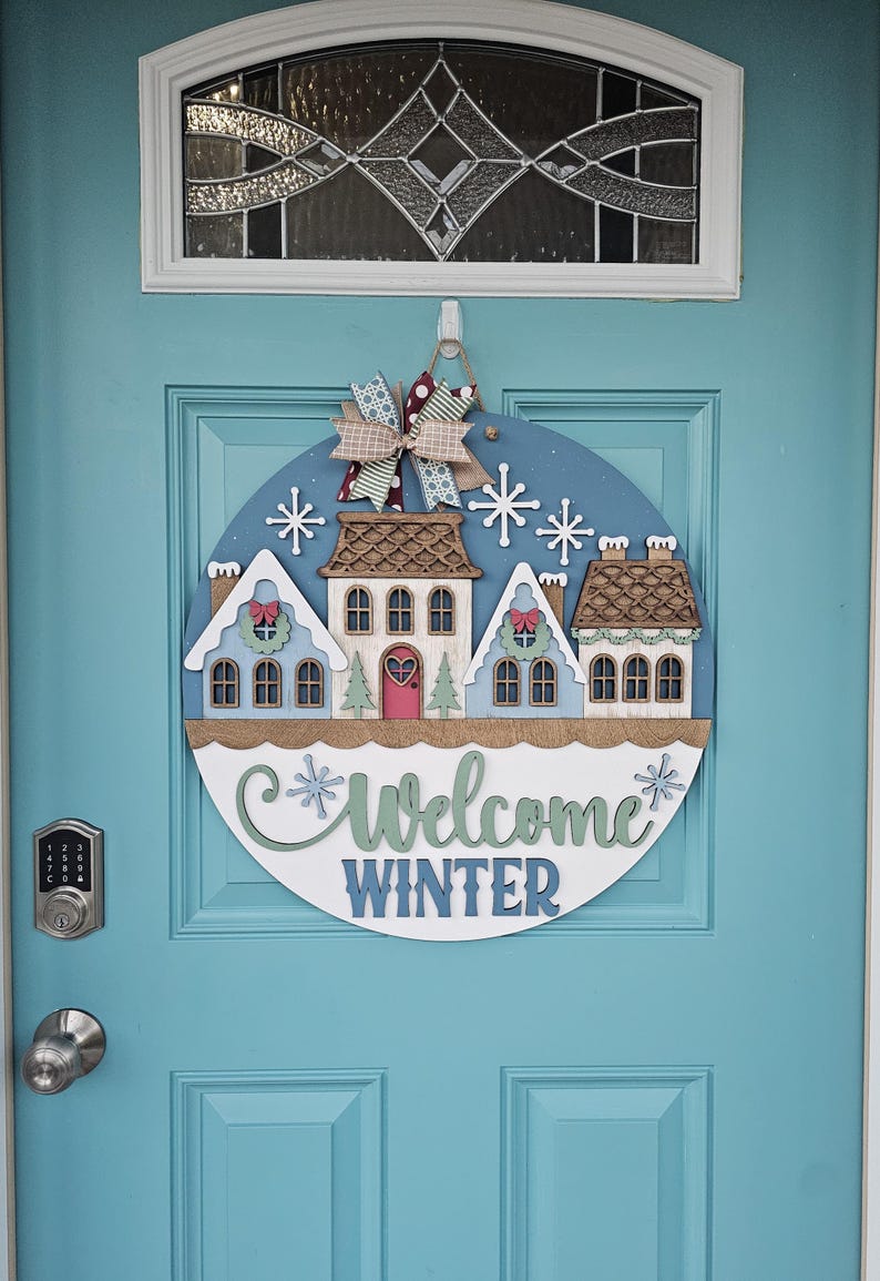 May include: A round wooden door sign with a winter theme. It features a blue background with white snowflakes and a row of houses. The sign says "Welcome Winter" in green and blue. A decorative bow hangs at the top.