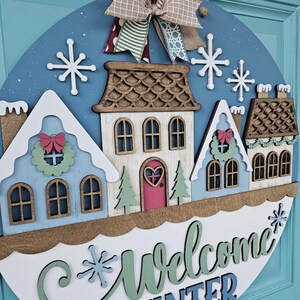 May include: A round wooden winter-themed door sign with a blue background and a decorative bow. The sign features a row of colorful houses, snowflakes, and the words "Welcome Winter" in script. The houses are painted in shades of blue, white, and brown.