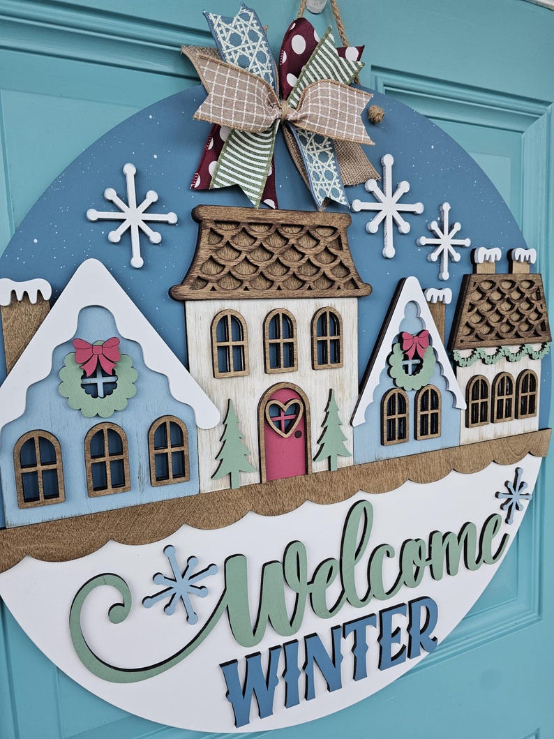 May include: A round wooden winter-themed door sign with a blue background and a decorative bow. The sign features a row of colorful houses, snowflakes, and the words "Welcome Winter" in script. The houses are painted in shades of blue, white, and brown.
