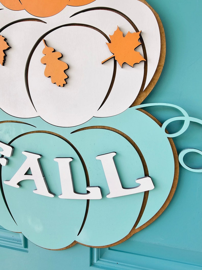 Op de afbeelding: Een houten herfstdeurdecoratie met twee witte pompoenen en een turquoise pompoen met het woord "FALL" in witte letters. De pompoenen hebben oranje bladeren bevestigd.
