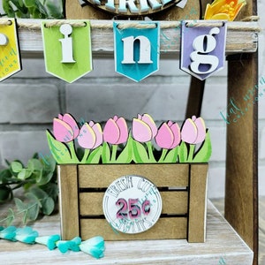 May include: A wooden crate with a sign that says "Fresh Cut Tulips 25&cent;" and a bunch of pink tulips. The crate is sitting on a wooden shelf with a banner that says "Spring" hanging above it.