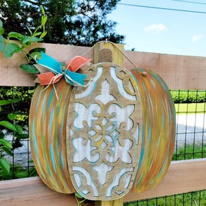 May include: A wooden pumpkin door hanger with a carved design and painted in shades of teal, orange, and yellow. The pumpkin is decorated with a teal and orange bow.
