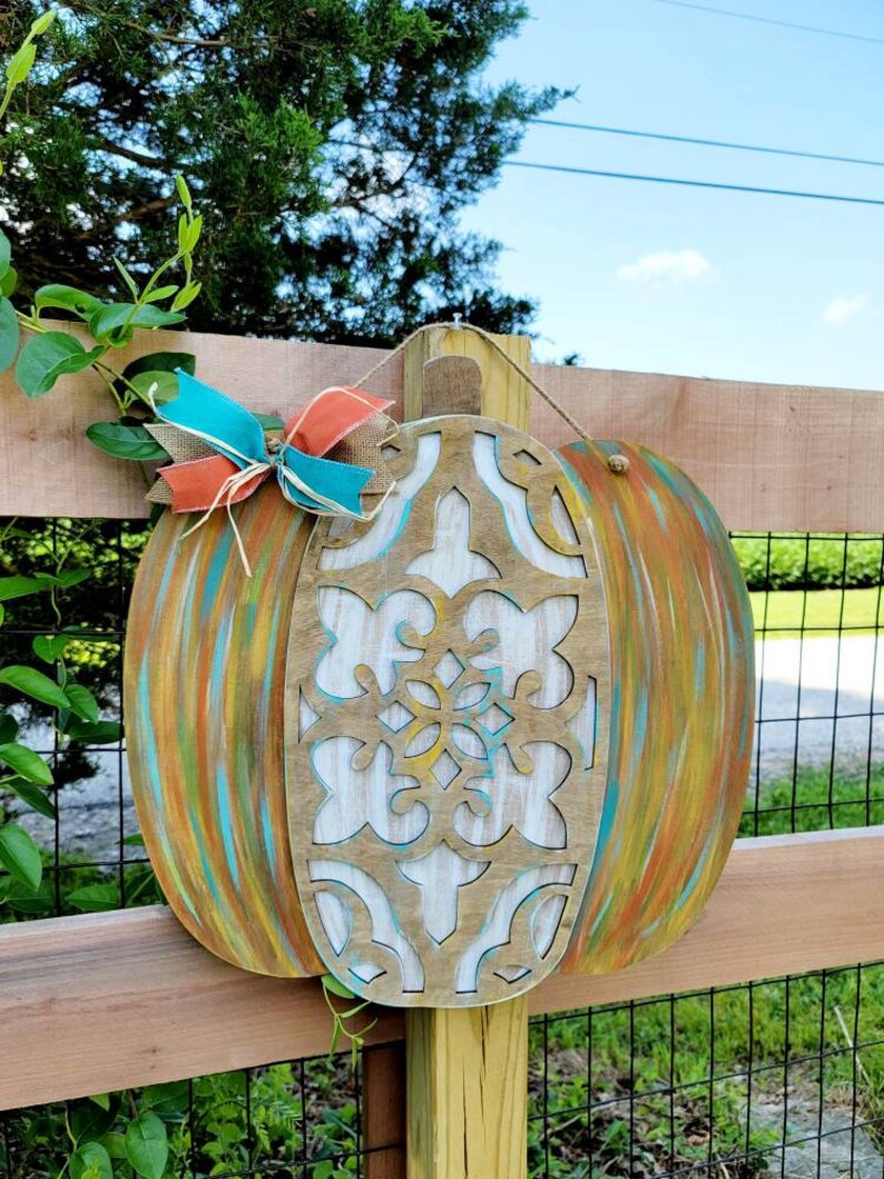 May include: A wooden pumpkin door hanger with a carved design and painted in shades of teal, orange, and yellow. The pumpkin is decorated with a teal and orange bow.
