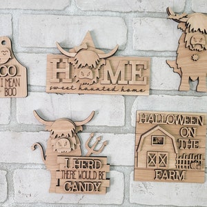 May include: Five wooden Halloween signs featuring a Highland cow design. The signs say "Moo I Mean Boo", "Home Sweet Haunted Home", "I Herd There Would Be Candy", "Halloween On The Farm", and a cow with horns and fangs.
