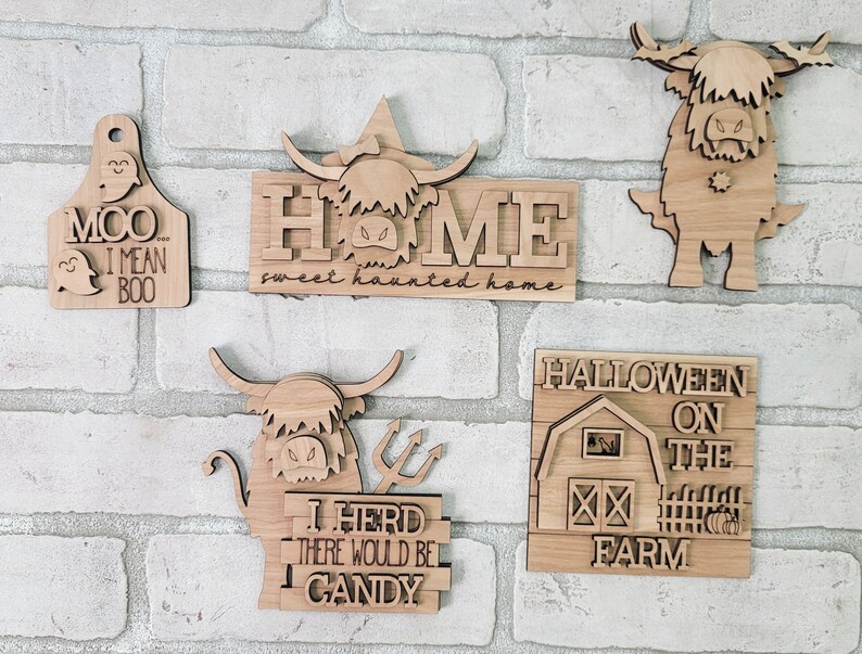 May include: Five wooden Halloween signs featuring a Highland cow design. The signs say "Moo I Mean Boo", "Home Sweet Haunted Home", "I Herd There Would Be Candy", "Halloween On The Farm", and a cow with horns and fangs.