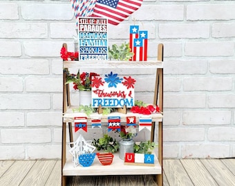 Pode incluir: Uma prateleira de madeira de dois níveis decorada com itens vermelho, branco e azul para o 4 de julho. A prateleira superior apresenta uma pilha de blocos de madeira com as palavras "4th of July", "Parades", "Fireworks", "Family", "Sparkler" e "Freedom". A prateleira inferior tem uma placa que diz "Fireworks & Freedom", uma guirlanda de bandeiras vermelho, branco e azul, um regador de metal e um bloco de madeira com as letras "U" e "A".