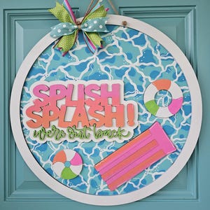 May include: A round, white-framed sign with a blue and white water pattern background. The sign features the words "SPLISH SPLASH! We're out back" in pink and orange lettering, along with a beach ball, a life preserver, and a pink pool float. A colorful bow tops the sign.
