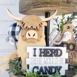 May include: A tiered wooden tray decorated with a brown and white highland cow figurine, a sign that reads "I herd there would be candy", and greenery.