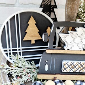 May include: A black and white tiered tray with a black truck carrying a Christmas tree, a black and white plaid Christmas tree, and a round black and white sign with a gold tree. The tray is decorated with a garland of black and white buffalo check beads and a sprig of frosted greenery.