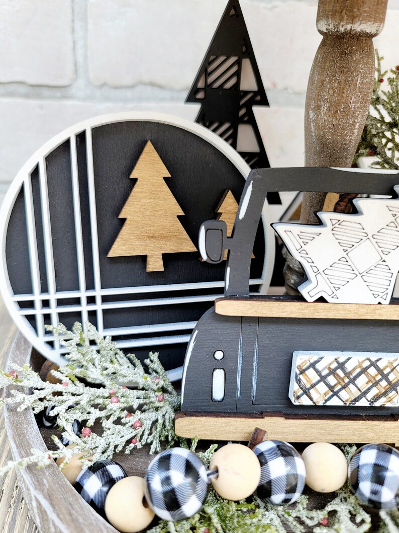 May include: A black and white tiered tray with a black truck carrying a Christmas tree, a black and white plaid Christmas tree, and a round black and white sign with a gold tree. The tray is decorated with a garland of black and white buffalo check beads and a sprig of frosted greenery.