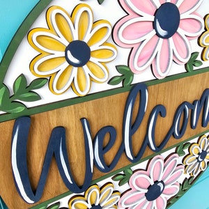 K&ouml;nnte beinhalten: Ein rundes Holzschild mit der Aufschrift "Willkommen" in Marineblau und einem floralen Design. Die Blumen sind rosa, gelb und wei&szlig; mit marineblauen Zentren.