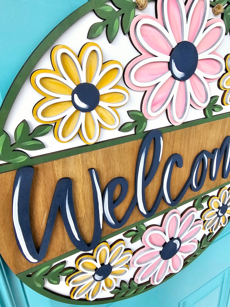 K&ouml;nnte beinhalten: Ein rundes Holzschild mit der Aufschrift "Willkommen" in Marineblau und einem floralen Design. Die Blumen sind rosa, gelb und wei&szlig; mit marineblauen Zentren.