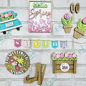 May include: A set of wooden spring-themed decorations, including a truck with flowers, a banner with the word "spring", a sign with the words "hello spring", a flower pot with flowers, a sign with the words "fresh flower market", a crate with flowers and the text "25c", and a pair of wooden legs.