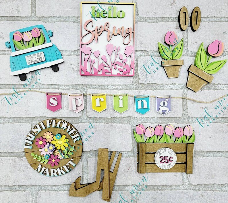 May include: A set of wooden spring-themed decorations, including a truck with flowers, a banner with the word "spring", a sign with the words "hello spring", a flower pot with flowers, a sign with the words "fresh flower market", a crate with flowers and the text "25c", and a pair of wooden legs.