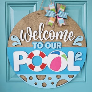 Puede incluir: Cartel de madera redondo con las palabras "Welcome to Our Pool" en blanco, con un fondo azul y un balón de playa y un salvavidas. Un lazo colorido está en la parte superior. El cartel está colgado en una puerta azul.