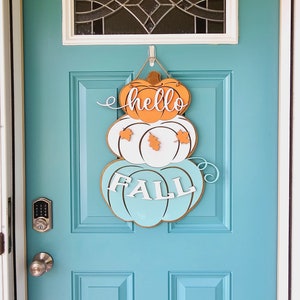 Op de afbeelding: Een turquoise voordeur met een houten pompoenkrans waarop "hello fall" staat in witte en oranje letters.